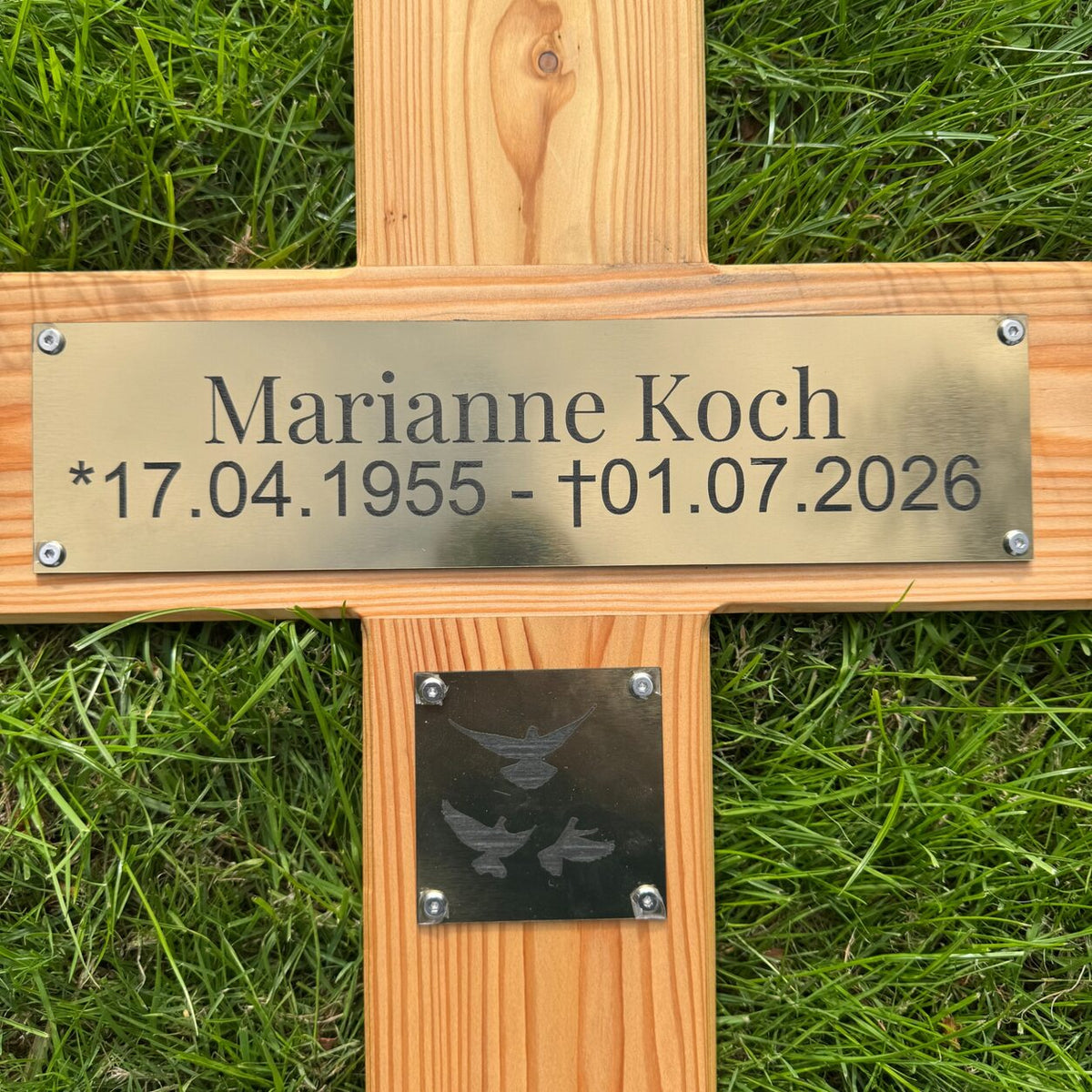 Nahaufnahme eines Holzgrabkreuzes mit goldfarbenem Schild, Namensgravur, Geburts- und Sterbedatum sowie Taubenmotiv
