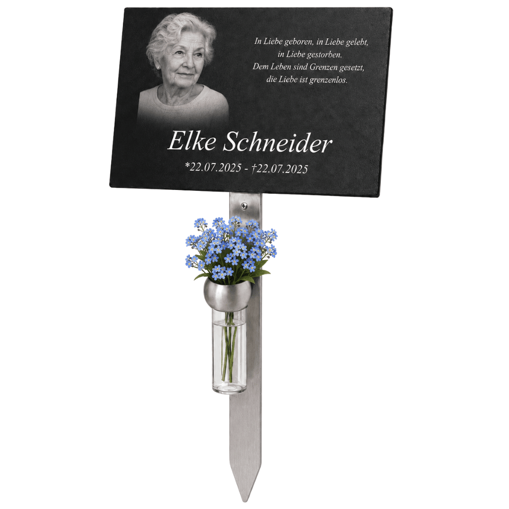 Schwarze Gedenktafel mit Porträt einer älteren Frau, persönlicher Inschrift und Namen ‚Elke Schneider‘ auf einem Metallstab. Darunter befindet sich eine kleine Vase mit blauen Blumen.