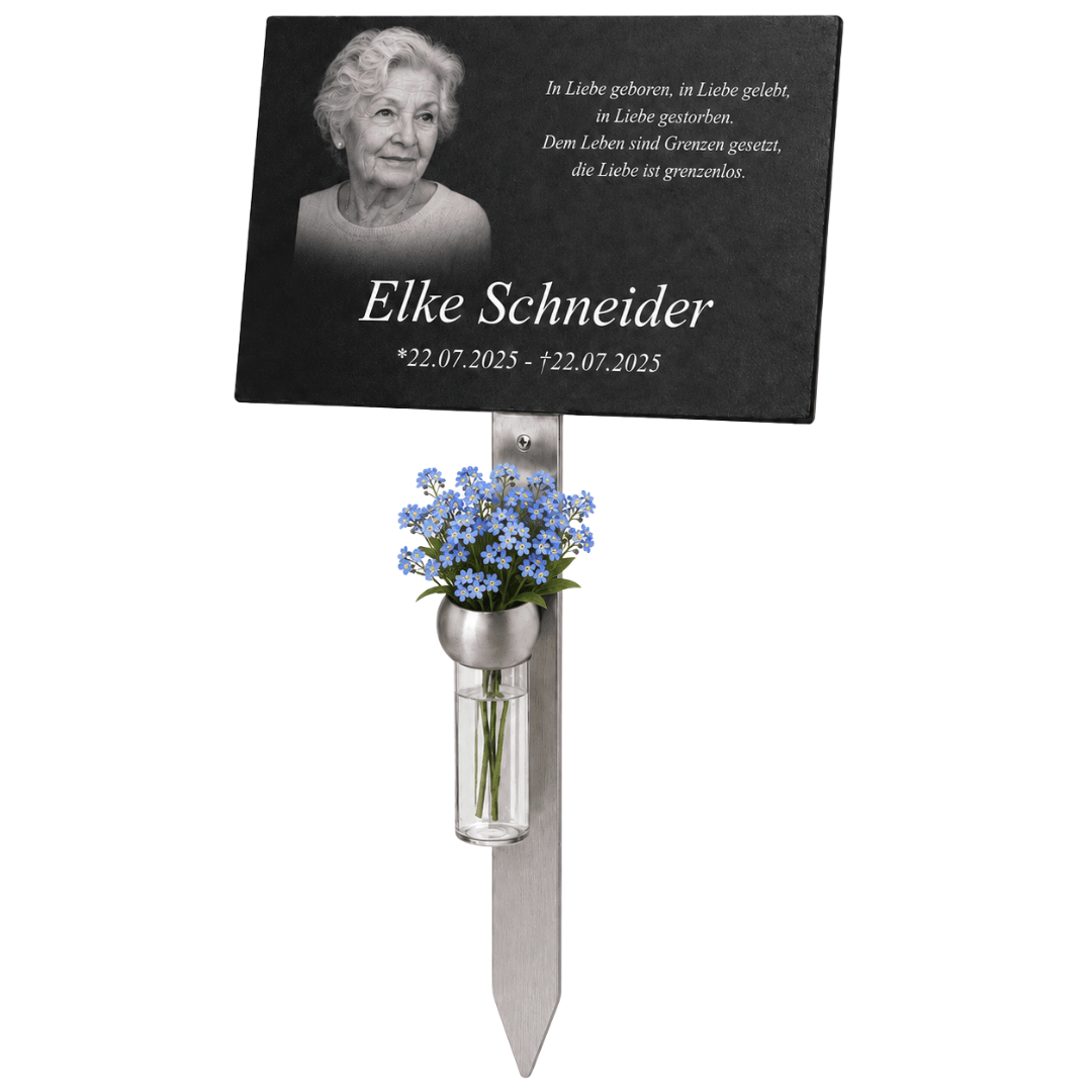 Schwarze Gedenktafel mit Porträt einer älteren Frau, persönlicher Inschrift und Namen ‚Elke Schneider‘ auf einem Metallstab. Darunter befindet sich eine kleine Vase mit blauen Blumen.