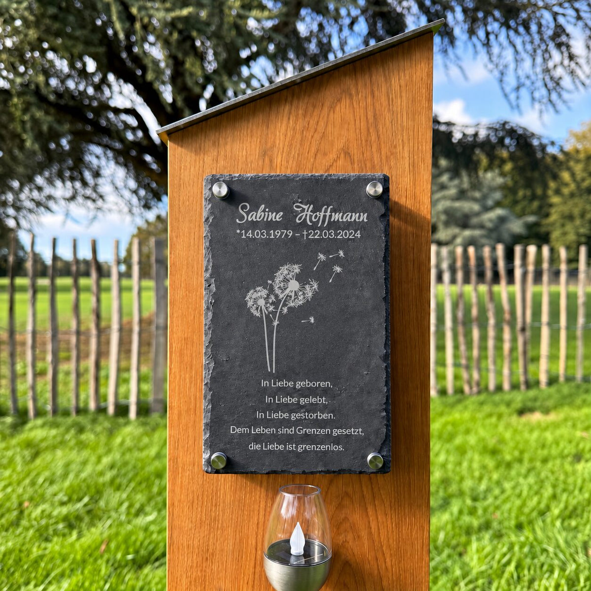 Nahaufnahme einer Schiefertafel an einer Holz-Grabstele mit Pusteblumenmotiv, Namen, Lebensdaten und Gedenkspruch.